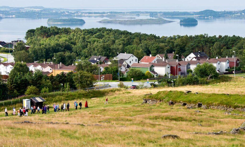 Bilde av flat-Jæren hvor Jernaldergården ligger, med hus, folk og gress i front og hav med øyer i bakgrunnen.