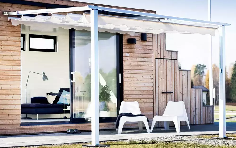 Hvit veggfestet-pergola på trefarget hus med terrassestoler, i solskinn med gress i front og bakgrunn.