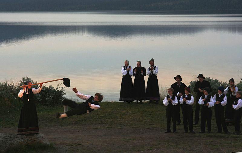 Bilde av voksne som danser og leker hallingkast og hallingdans i solnedgang. Ikledd bunader og ved et blikkstille vann. Vi tipser om 17 morsomme 17. mai-leker for voksne og barn.