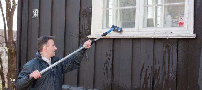 Mann som vasker brunt hus med kost og forlengerskaft til husvask.