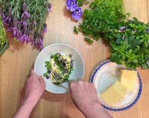 Bilde av blomstersmør pyntet med blomster. En hand blander sammen ingrediensene til smøret med en gaffel i en hvit skål på et bord med blomster, urter og en skål med romtemperert smør. Dette smøret imponerer på både 17. mai, i bryllup, dåp eller konfirmasjon.