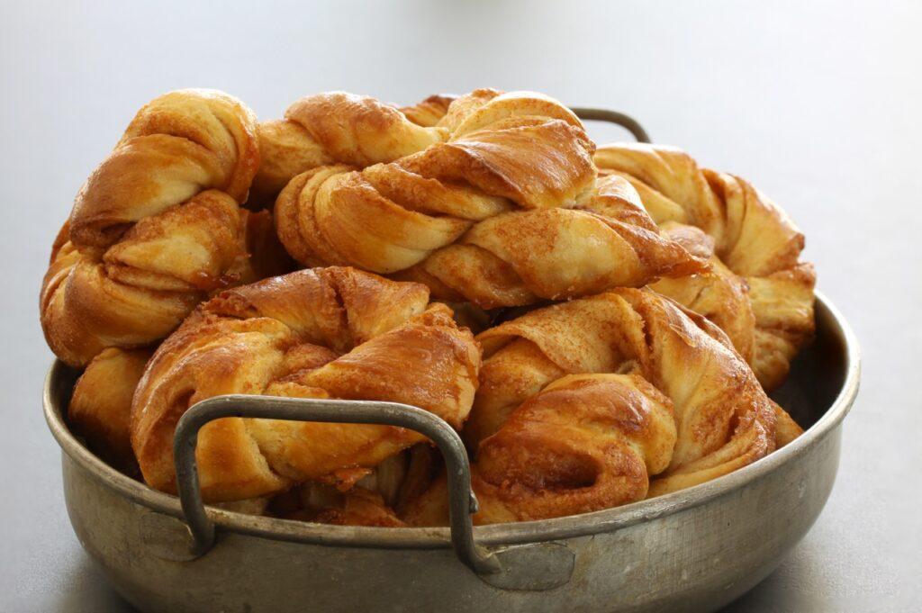 Bildet viser en stabel med gyllenbrune kanelknuter, som er en type kanelboller, perfekt stekt med et fristende lag av sukker og kanel. Disse ferske bakverkene ser svært innbydende ut, med den krøllete formen som gir en ekstra deilig tekstur. Kanelboller som dette er en perfekt måte å feire Kanelbollens dag den 4. oktober. Dagen er en ypperlig anledning til å nyte en klassisk nordisk favoritt som varmer både smaksløkene og sjelen på en høstdag.