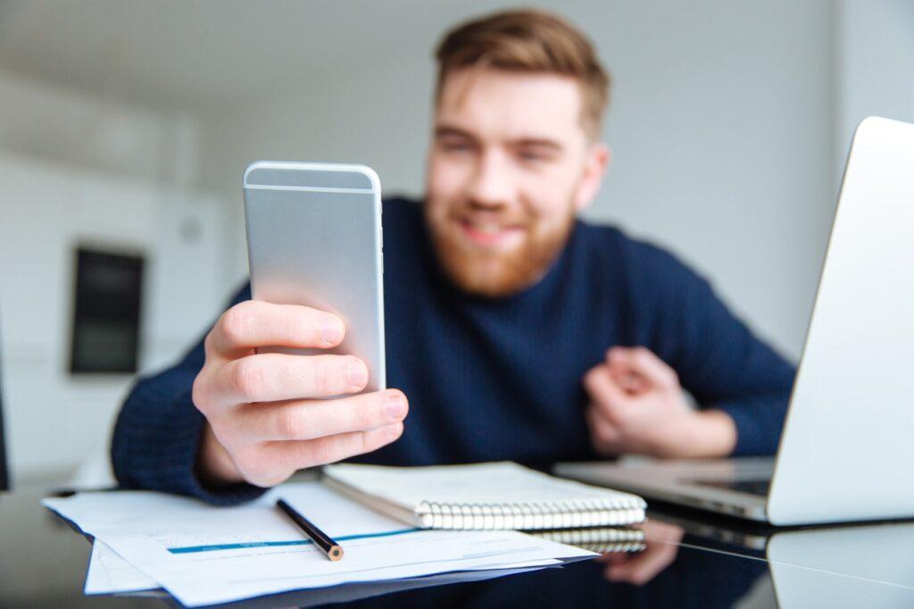 Man som er fornøyd fordi han sparer oenger ved å føre budsjett. På pult med ark, PC, smilende med en mobil i handen.