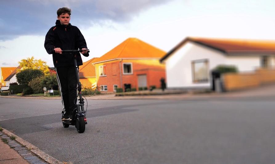 Ung gutt som kjører en el-sparkesykkel, hus i bakgrunnen. Vi forteller deg alt du må vite når du skal kjøpe el-sparkesykkel til barn.