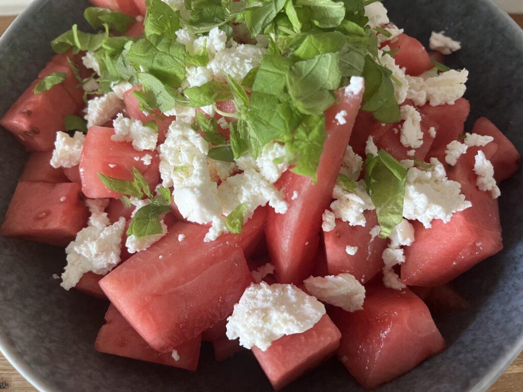 Bilde av frisk, enkel, sunn og kjapp sommersalat med vannmelon, fetaost og mynte på et bord i en blå skål. Vi gir deg oppskrift på det smakfulle tilbehøret som passer ypperlig til sommerens grilling.
