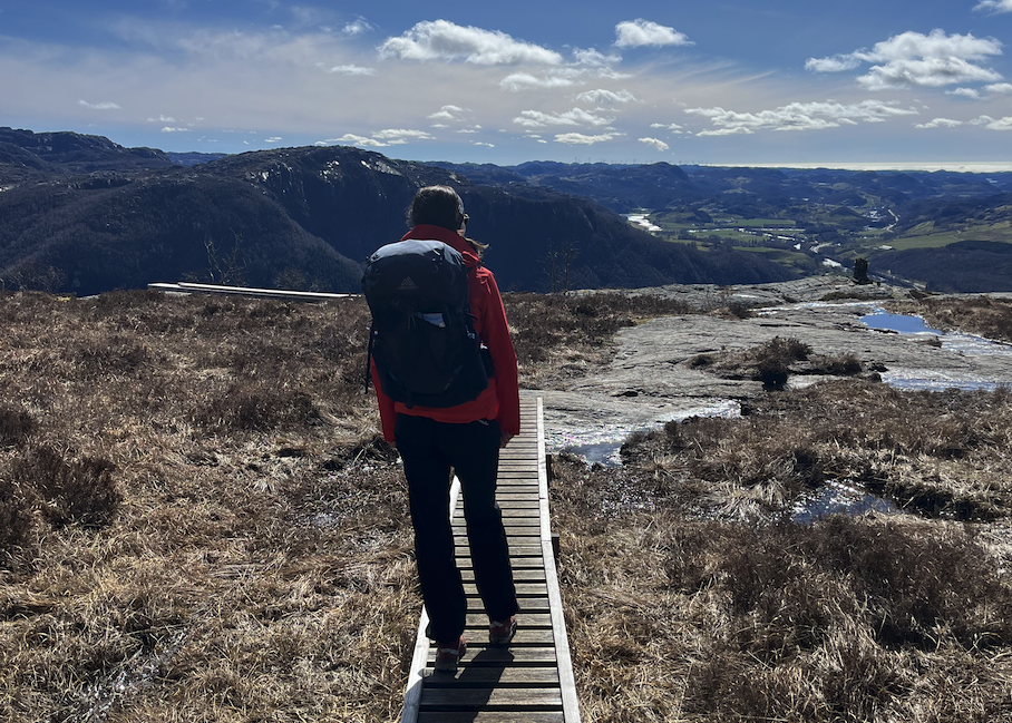 En person som står på i en skog, foran et tjern, med utsikt over skog og vakker sommer-kledd natur. Damen har på seg en grå tursekk, en blå treningstights, caps og en svart genser. Vi har fått eksperthjelp til å lage en guide på hvordan man velger riktig tursekk, som liten tursekk, mellomstor eller stor. I tillegg forklarer vi forskjellene på tursekk dame vs tursekk til mann, og legger fram populære sekker fra Osprey og Bergans.