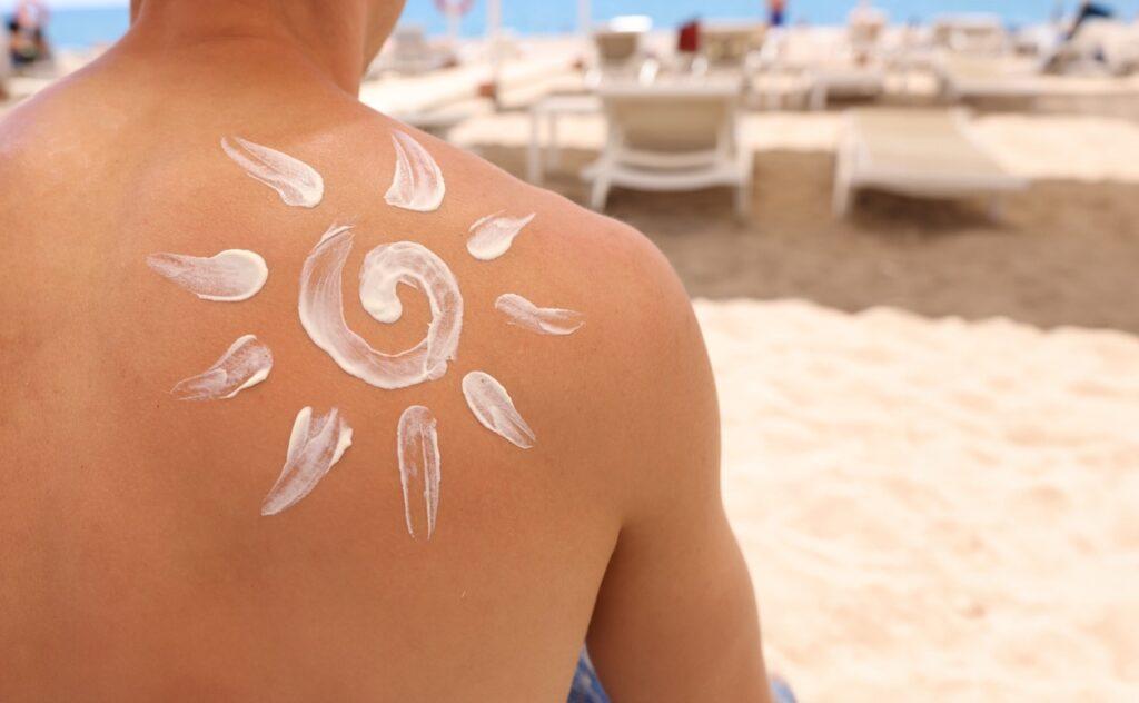 Bilde av en person som sitter med ryggen til på en strand. På ryggen er det laget en sol av solkrem for å indikere at personen smører seg med solkrem før hen skal sole seg. Vi svarer på spørsmål som: Hva er UV-indeks? Hvordan sjekke UV-indeks? Når er UV-indeksen høyest? Hva er høy UV-indeks? og hvilken UV-indeks blir man brun av?