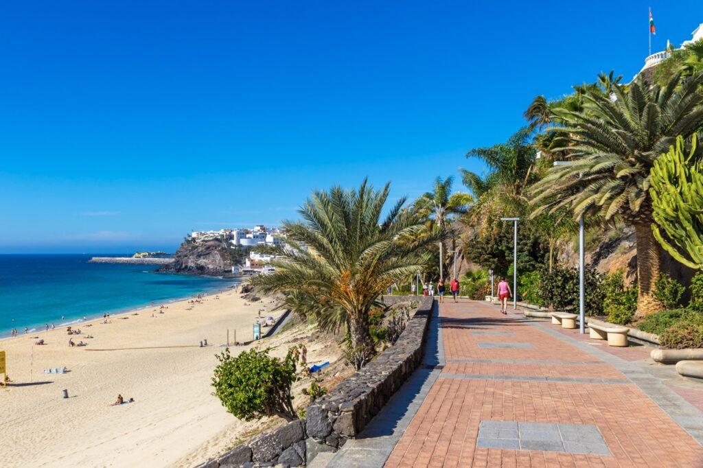 Bilde av en vakker strand på Fuerteventura, Kanariøyene. gylden strand, blått hav og blå himmel. På høyre side går en promenade langs stranden. Her finner du pakkereiser til kanariøyene. Finn restplasser til blant annet Gran Canaria, Tenerife, Lanzarote og Fuerteventura. 