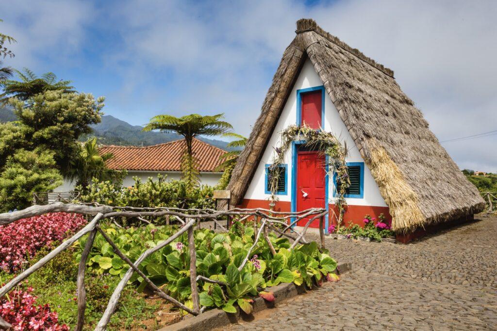 Bilde av et tradisjonelle trekantede hus med stråtak. Huset er av hvit mur med røde og blå detaljer. Her finner restplasser til Madeira i Portugal slik at du enkelt kan finne billig reise til den vakre øya.