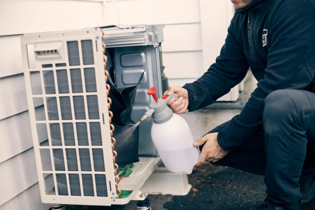 Bilde av en installatør som har service på varmepumpe, utedelen som henger lavt nede på husveggen. Vi har snakket med eksperten som forteller alt du må vite om service på varmepumper, uavhengig av om du har luft til luft-varmepumpe fra Mitsubishi, Toshiba, Daikin eller Panasonic.