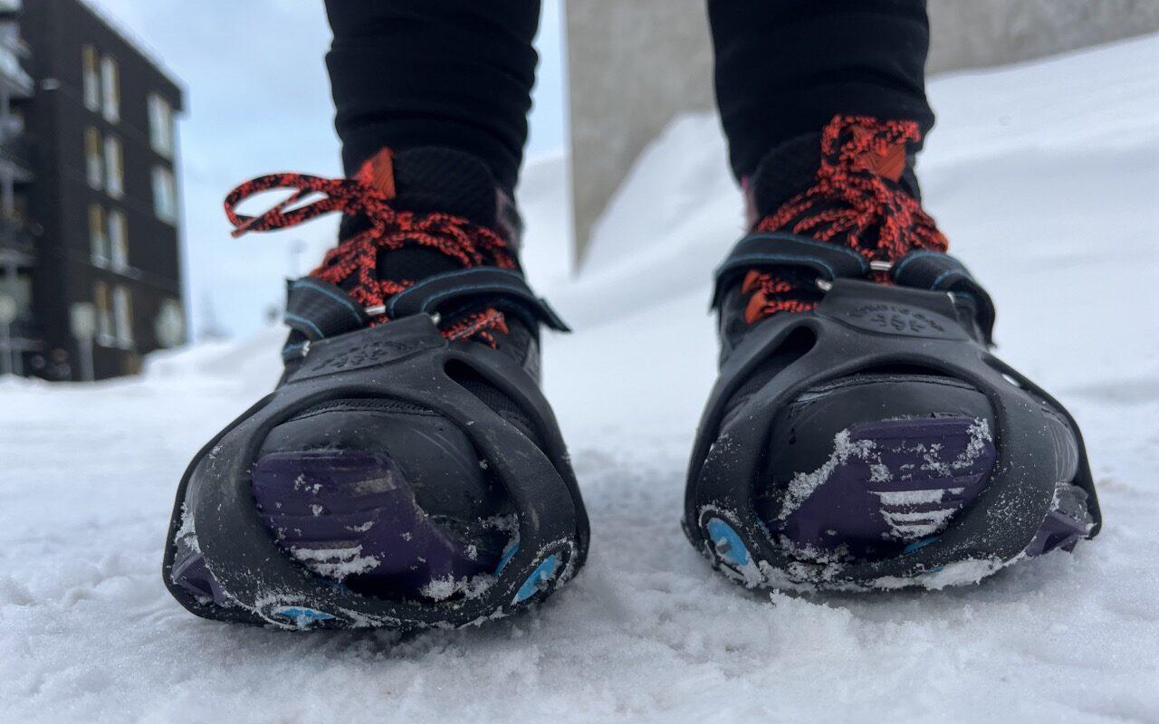 Bilde av person som står ute i naturen med hus og blokker i bakrgunnen og som har på seg Nordic Grip-brodder av typen Nordic Grip running. Broddene er tredd utenpå et par med svarte joggesko fra Asics med oransje detaljer og lisser og personen står på et snødekket underlag. Vi forteller hvilke brodder som er best til ulike formål!