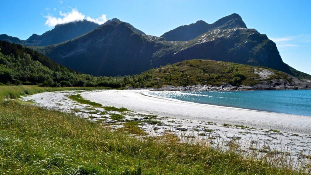 Bilde av en hvit strand med blått hav og fjell i bakgrunnen på Kjerringøy. Her finner du overnatting på Kjerringøy, blant annet Kjerringøy Bryggehotell som har blitt brukt i innspillingen til Hver gang vi møtes. Du finner også Kjerringøy rorbusenter og Kjerringøy Camping.