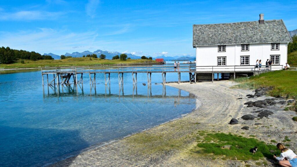 Bilde av det gamle handelsstedet på Kjerringøy. Rundt er det turkis hav. Her finner du overnatting på Kjerringøy, blant annet Kjerringøy Bryggehotell som har blitt brukt i innspillingen til Hver gang vi møtes. Du finner også Kjerringøy rorbusenter og Kjerringøy Camping.