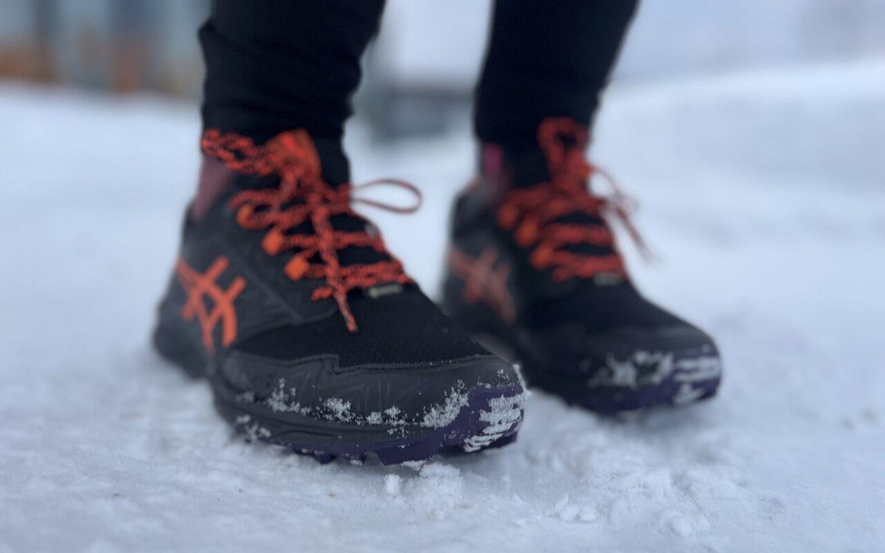 Bilde av person med asics-piggsko på føttene. Personen står med de svarte skoene med oransje lisser og detaljer ute i snø i et nabolag. Vo forteller hvor du kan finne billige piggsko på tilbud!