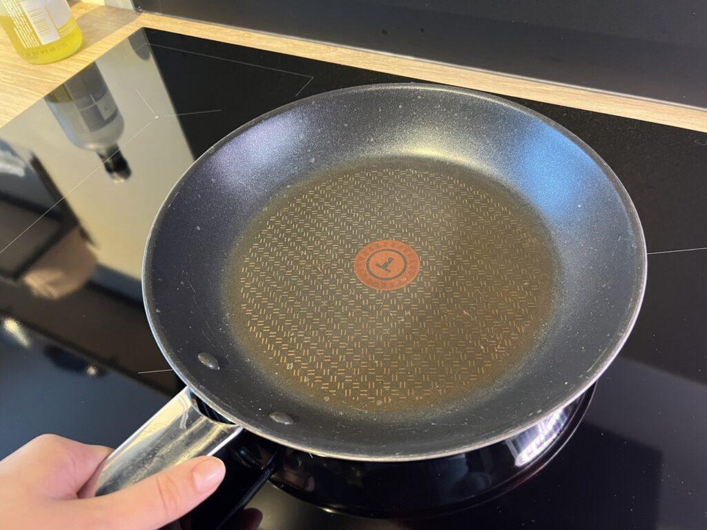 Bildet viser en svart Tefal-stekepanne med et karakteristisk rødt «Thermo-Spot»-merke i midten. Pannen har et diskret, gyllent rutemønster rundt det røde feltet og viser tegn til bruk i form av lys misfarging. Den ligger på en blank, svart induksjonskoketopp, og man kan skimte en flaske i speilingen til venstre i bildet. Nederst i venstre hjørne holder en hånd i panne-håndtaket, som er i blankt metall. Bakgrunnen består av en del av kjøkkenbenken.Produsenter av non-stick stekepanner får kritikk.