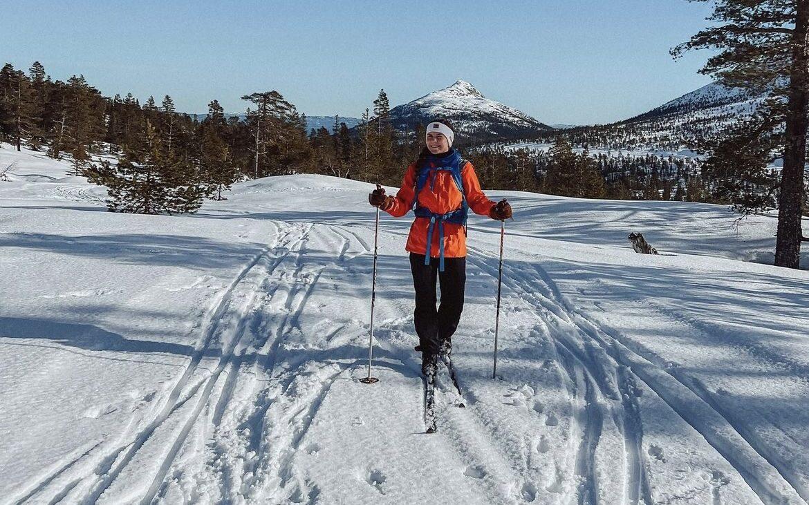 Bilde av Runa Sommerfelt Ovenstad på skitur med langrenn i et snødekket landskap i Norge. Hun smiler mot kamera og har vakre snødekkede fjell og skog i bakgrunnen. Vi har snakket med Runa og ville høre hva hun mener man må pakke med seg i tursekken på toppturer om vinteren. Hun deler både sin egen pakkeliste og sine favoritt-toppturer i Norge.