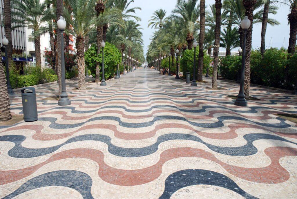 Bilde av den kjente promenaden i Alicante, som ligger langs Costa Blanca i Spania, hvor du kan se den bølgende mosaikken på bakken i fargende rød, hvit, og mørk blå. På siden er det grønne trær som strekker seg opp mot himmelen. Her får du tips til hva du må se og gjøre i Alicante, som blant annet besøke Santa Bárbara-slottet, Basilica de Santa Maria, Tabarca-øya, Mercado Central og Explanada de España.