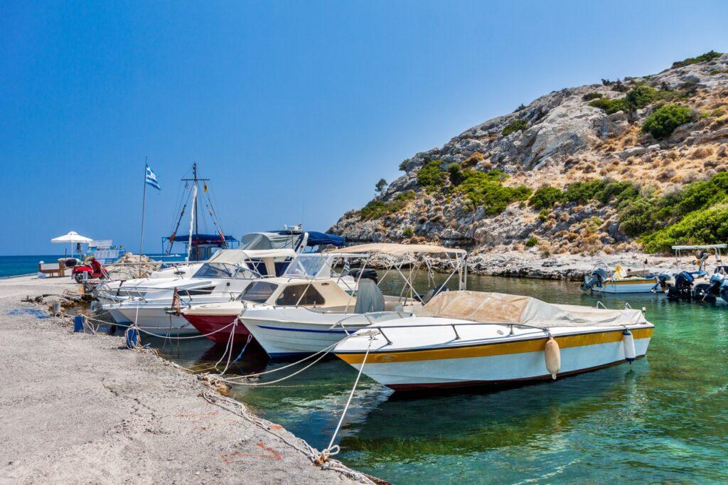 Et bilde av en liten havn på Rhodos, Hellas, med flere småbåter fortøyd ved en brygge. En gresk flagg vaier i vinden ved bryggen, og en person kan sees sittende under en parasoll i bakgrunnen. Området er omgitt av steinete åser med grønn vegetasjon. Vannet er klart og speiler den blå himmelen over. Rhodos i Hellas er en drøm, og her får du både billige reiser og pakkereiser til den greske øya. Vi gir deg også fem ting du må se og gjøre på Rhodos, samt svar på ofte stilte spørsmål om øya.