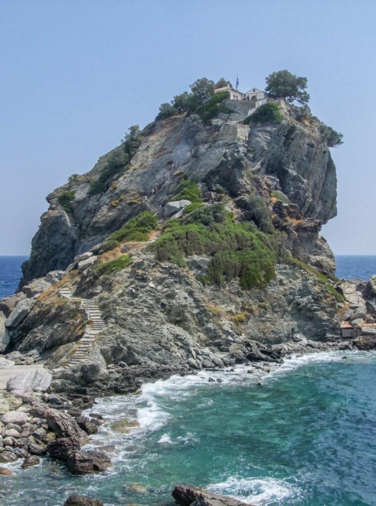 Bilde av en klippe i havet som har en smal steinsti til fastlandet. På klippen er en lang steintrapp og på toppen kan man skimte en kirke. Havet rundt er blått og slår mot klippene. Vi hjelper deg med å finne billige reiser til Skopelos som er den greske øya hvor «Mamma Mia» ble spilt inn. Vi gir deg også fem tips til hva du må se på øya som ligger i Hellas, som blant annet Agios Ioannis og Cape Amarandos.