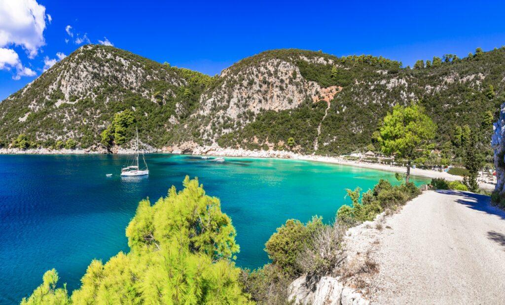 Knallblått hav omringet av grønn natur og en hvit strand. På havet kan man også se en seilbåt. Vi hjelper deg med å finne billige reiser til Skopelos som er den greske øya hvor «Mamma Mia» ble spilt inn. Vi gir deg også fem tips til hva du må se på øya som ligger i Hellas, som blant annet Agios Ioannis og Cape Amarandos.