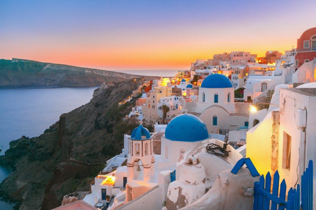 Utsiktsbilde fra byen Oia på Santorini som soler seg i en spektakulær solnedgang. På høyre siden kan man se de velkjente hvite murhusene med blå detaljer, og på venstre side har man flott utsikt over Egeerhavet. Vi har skrevet om fem ting man bør se og gjøre på Santorini i Hellas, samt avslører hvilket hotell på Santorini som er kåret til det beste. Her finner du også restplasser til Santorini og billige reiser til de greske øya