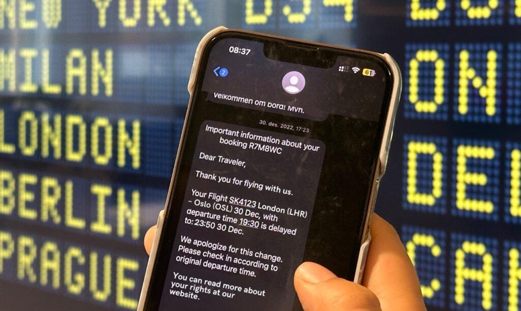 Person som holder opp en telefonen hvor man ser en melding fra flyselskapet om at flyet er forsinket. I bakgrunnen kan man se en tavle hvor flyreisene vises på flyplassen. Vi har snakket med Forbrukerrådet som forteller alt man må vite om standardkompensasjon når man skal ut å fly og det er forsinket fly.