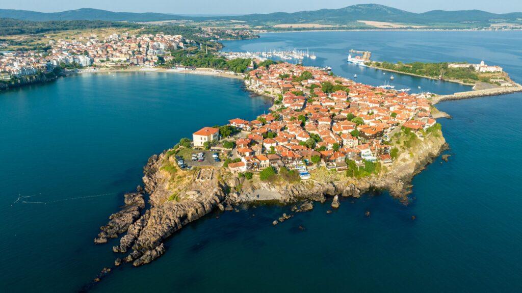 Bildet viser en spektakulær luftfoto av Sozopol i Bulgaria som er dekket av tettbygde hus med røde tak. Halvøyen strekker seg ut i et stille, blått hav og er omringet av klipper og små steiner i vannkanten. Vi hjelper deg med å finne billig ferie, og svarer på spørsmål som hvor er det billig å reise, hva koster en halvliter øl, og avslører fem land som er kåret til de billigste i Europa for nordmenn å feriere i.