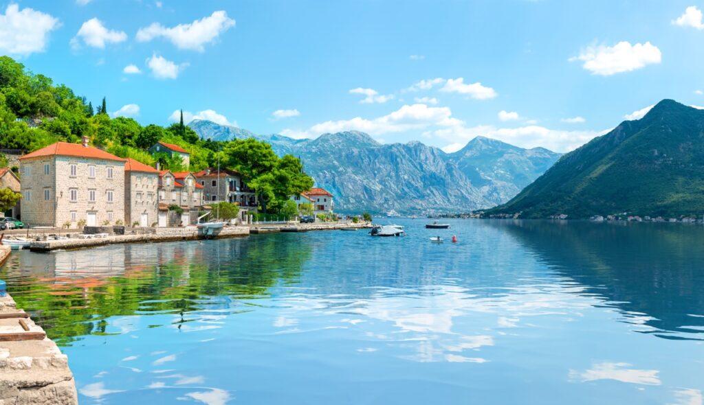 Bildet viser byen Kotor i Montenegro. Landsbyen består av gamle steinhus med røde tak som ligger tett i tett langs en rolig havnefront. Små båter flyter på det speilblanke vannet, som reflekterer en klar blå himmel og de omkringliggende grønne åsene. Bakgrunnen domineres av dramatiske fjelltopper som skaper en imponerende og vakker naturskjønn ramme rundt denne sjarmerende kystlinjen. Vi hjelper deg med å finne billig ferie, og svarer på spørsmål som hvor er det billig å reise, hva koster en halvliter øl, og avslører fem land som er kåret til de billigste i Europa for nordmenn å feriere i.