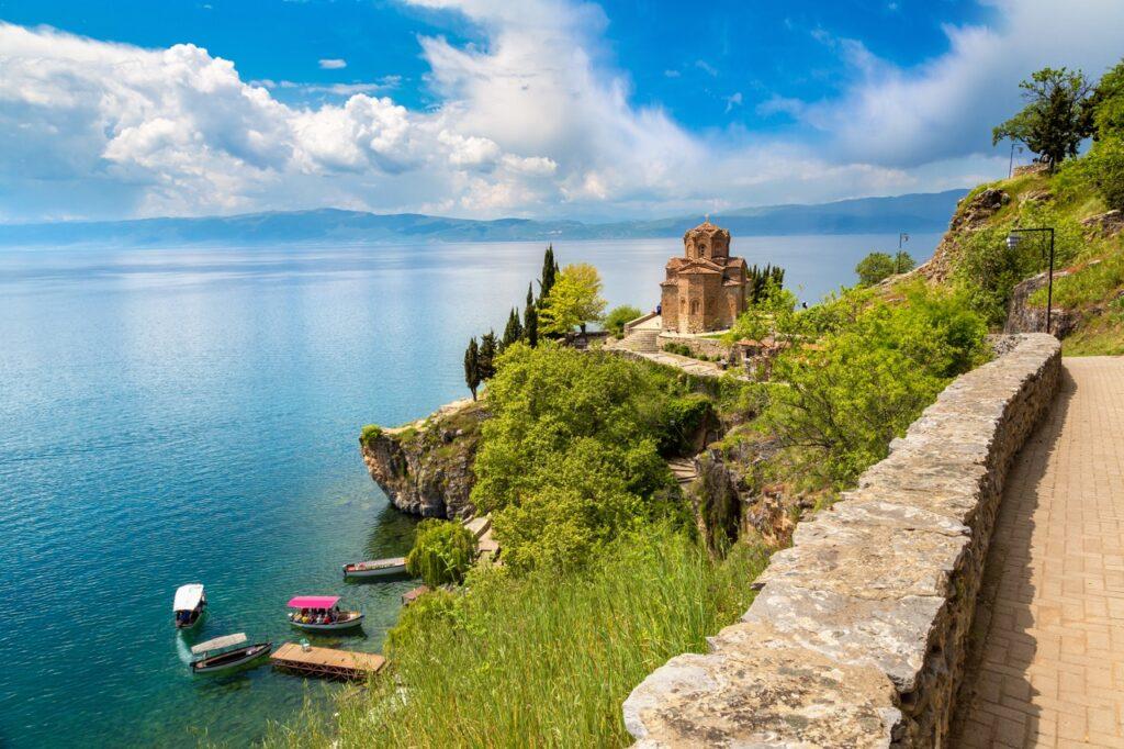 Bildet viser en pittoresk utsikt over Jovan Kaneo-kirken i Nord-Makedonia bygget på en klippe ved bredden av vakre Ohridsjøen. Kirken, som er laget av rødbrune steiner med detaljerte arkitektoniske utsmykninger, omgis av frodige grønne trær og busker. Foran kirken ligger en steinbelagt sti som fører langs klippens kant, og noen små båter er fortøyd i det klare blå vannet nedenfor. I bakgrunnen kan man se et fjellandskap og en dramatisk himmel med spredte skyer. Vi hjelper deg med å finne billig ferie, og svarer på spørsmål som hvor er det billig å reise, hva koster en halvliter øl, og avslører fem land som er kåret til de billigste i Europa for nordmenn å feriere i.