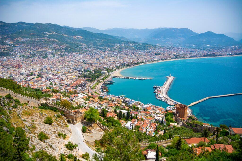 Bildet viser en flott utsikt over Alanya i Tyrkia, med gamle steinmurer og det historiske vakttårnet, Det røde tårn, i forgrunnen. Byen er tett befolket med fargerike hus som strekker seg ned mot en lang sandstrand og en travel havn. Båter ligger forankret i det azurblå vannet, som strekker seg mot fjell i det fjerne under en klar blå himmel. Hele scenen er omringet av frodig grønne fjell og gir et inntrykk av en pulserende by som lever i harmoni med sin rike historie og naturskjønne omgivelser. Vi hjelper deg med å finne billig ferie, og svarer på spørsmål som hvor er det billig å reise, hva koster en halvliter øl, og avslører fem land som er kåret til de billigste i Europa for nordmenn å feriere i.