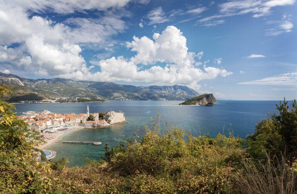 utsikt over Budva, Montenegro, sett fra en høyde. Byen ligger ved kysten og er omkranset av høye fjell i bakgrunnen. Gamlebyen med sine lyse bygninger og et prominent kirketårn ligger ved en liten halvøy, flankert av en stor strand og det krystallklare Adriaterhavet. Montenegro er et spennende land langs Middelhavet som er rangert til å være et billig land for nordmenn å feriere i. Vi hjelper deg med å finne pakkereiser som inkluderer fly og hotell, samt restplasser til Montenegro.