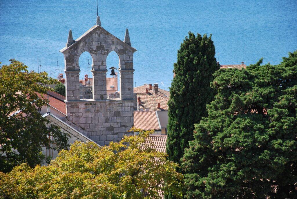 En stemningsfull utsikt over en historisk kirkeklokketårn i Pula, med to synlige klokker, bygget i stein og flankert av spisse bueåpninger. Tårnet er omgitt av frodige trær og hustak, med glimt av en rolig blå sjø i bakgrunnen. Vi hjelper deg med å finne riktig destinasjon til din Kroatia-ferie, og har snakket med Gjermund Glesnes, som står bak reisebloggen Detvondeliv.no, som forteller om populære destinasjoner som Brac, Hvar, Split og Dubrovnik.