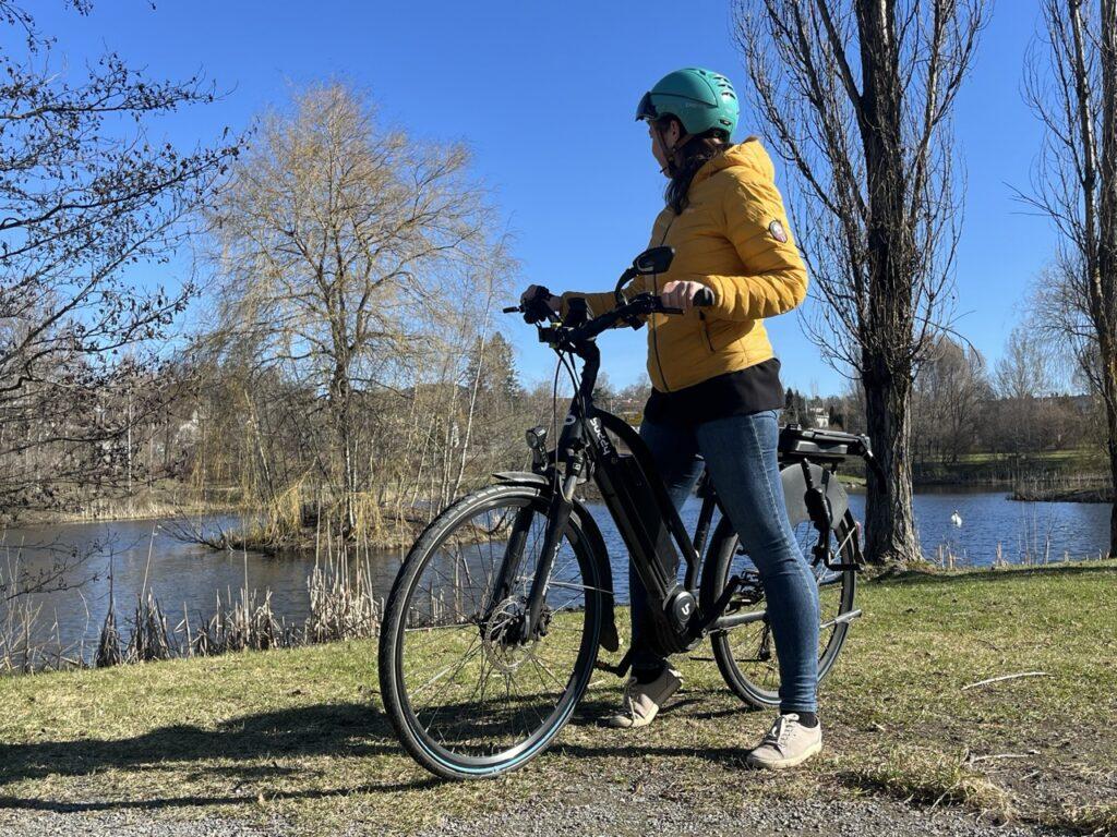 Bilde av dame som står ved en elsykkel til dame fra Buddy Bike. Hun har på seg en gul dunjakke, dongeribukser og en blå sykkelhjelm og står med elsykkelen sin på en gressplen med et vann i bakgrunnen. Vi har snakket med sykkeleksperten som forteller hvordan du velger riktig sykkel. Han forteller når du bør velge en terrengsykkel, når du bør velge hardtail eller fulldempet, og når det passer best med en landeveissykkel, gravelsykkel, vanlig sykkel, lastesykkel eller elsykkel. Han gir også et supertips for hvordan du kan få to sykler i én.