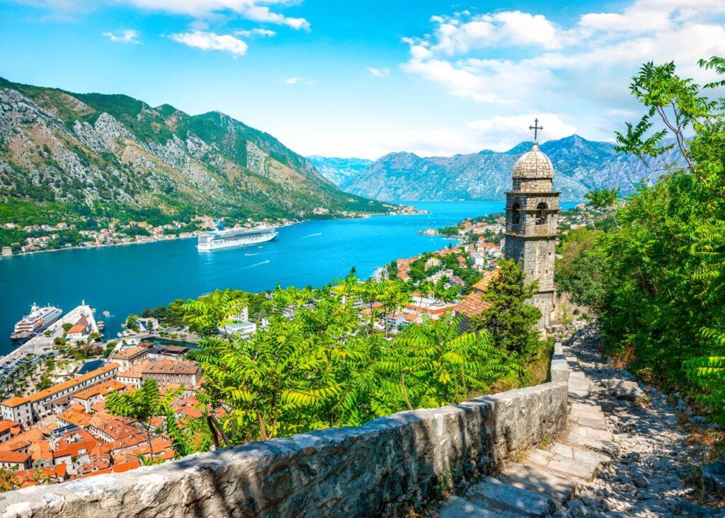 Utsikt over den gamle bydelen i Kotor, Montenegro, fra en høyde med en steinvei og et gammelt steintårn i forgrunnen. Bakgrunnen viser en klar, solrik dag med blå himmel og flere fjell som omslutter en bukt hvor cruiseskip og båter er ankret opp. Montenegro er et spennende land langs Middelhavet som er rangert til å være et billig land for nordmenn å feriere i. Vi hjelper deg med å finne pakkereiser som inkluderer fly og hotell, samt restplasser til Montenegro.