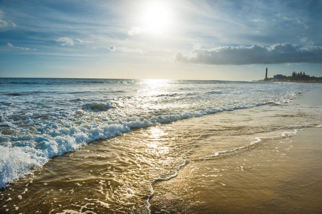 Bilde av en gylden strand i Maspalomas på Gran Canaria. Solen skinner og reflekteres i det klare vannet. I bakgrunnen ser man et fyrtårn. 