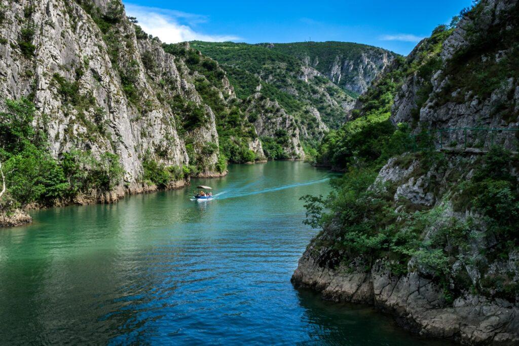 Et bilde av Matka Canyon i Nord-Makedonia, der en liten båt glir stille gjennom det turkisfargede vannet som er omringet av bratte klipper og frodig, grønn vegetasjon. Høye fjellvegger dominerer landskapet. Vannet reflekterer de lysegrønne tonene fra trærne og det mørkeblå fra himmelen. Nord-Makedonia har blitt rangert som landet i Europa som er billigst for nordmenn å feriere i. Vi har samlet fem ting du bør se og gjøre i landet, samt hjelper deg med å finne billige reiser ved hjelp av restplasser.