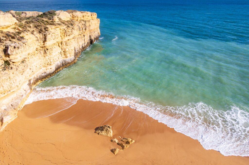 Bildet viser en vakker strand i Albufeira langs Algarve-kysten i Portugal. Stranden består av gyllen sand med noen få spredte steiner, og ligger ved foten av høye, klippeformasjoner av lyse steiner. Bølgene skvulper forsiktig inn mot kysten fra det klare, blågrønne vannet. Havet strekker seg ut i horisonten under en klar, blå himmel, og gir et fredelig og innbydende inntrykk. Vi hjelper deg med å planlegge ferien til Algarve-kysten i Portugal. Vi gir deg alt fra tips til byer; Lagos, Faro, Tavira, Albufeira og Sagres. Samt hvor du kan finne hotell langs Algarve-kysten og shoppingmulighetene.