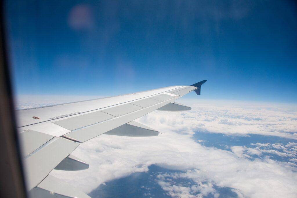 Flyfoto tatt fra et vindu, med utsikt over flyets vinge og skyene under. Himmelen er klar og blå, med noen skyer spredt i horisonten. Vi hjelper deg med å finne billige reiser med blant annet restplass-fly. Vi viser til fem tjenester som lar deg enkelt finne billige flybilletter; Apollo, Momondo, Restplass.no, Skyscanner og FINN reiser