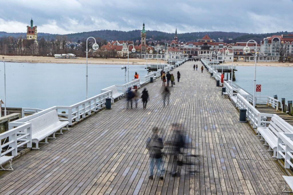 Bilde av den 500 meter lange tremoloen i Sopot, Polen. På moloen går det flere folk som får en flott utsikt over sjøen. I bakgrunnen på bildet kan man se byen Sopot. Sopot i Polen er perfekt for deg som liker å bade eller å nyte luksus på ett av spaene. Sopot har flere hotell som passer forskjellige budsjetter enten du er ute etter luksus eller budsjettvennlige hotell i Sopot. Her finner du billig ferie til sol og varme.