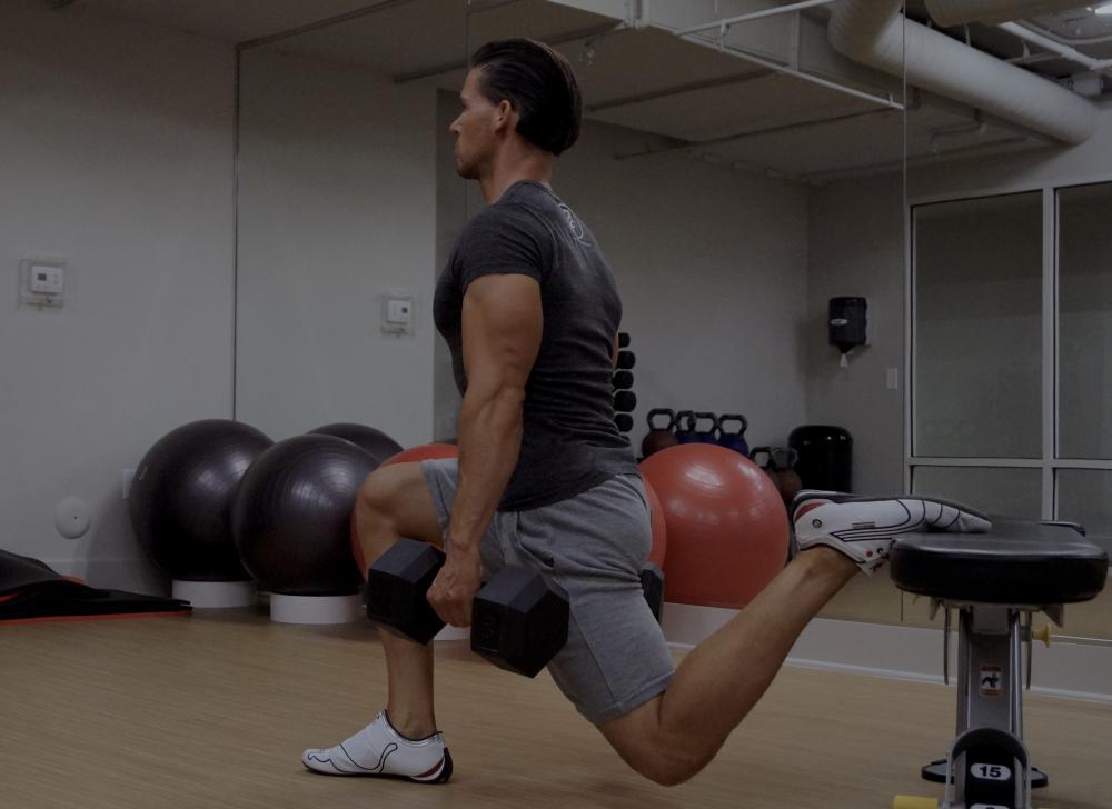 Bilde av personlig trener Cornelis Elander som utfører en øvelse kjent som "Bulgarian split squat" i et treningsstudio. Han holder en manual i hver hånd, og det ene beinet er plassert bak ham på en benk, mens det andre beinet er bøyd foran ham. Han står i en treningsrom med speilvegger, hvor vi kan se refleksjonen hans, og bak ham er det flere treningsballer og annet treningsutstyr. Han har på seg en mørk t-skjorte, grå shorts og hvite treningssko. Øvelsen fokuserer på å trene benmuskulaturen, spesielt quadriceps, hamstrings og gluteus. Dette er en styrkeøvelse med fordeler for bein og rumpe, og en av styrkeøvelsene i vårt 30-dagers gratis treningsprogram og styrkeprogram.