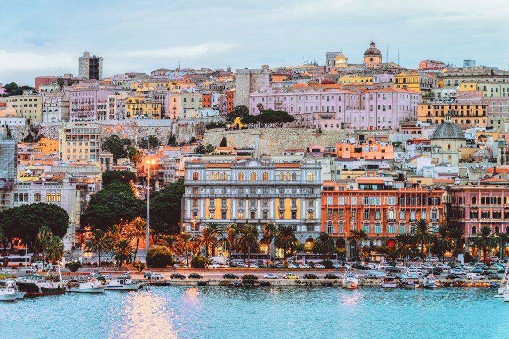 Et fargerikt bilde av byen Cagliari på Sardinia, Italia. Bildet viser en rekke bygninger i pastellfarger som ligger tett sammen på en høyde, med noen eldre strukturer og tårn som stikker opp. Ved foten av bakken er det en rekke palmer og en travel havnepromenade med parkerte biler og båter fortøyd ved kai. Lyset fra gatelykter og vinduer gir en varm glød. Middelhavsøya Sardinia byr på både luksus og spennende historie, og her finner du krystallklar sjø og kritthvite strender. Vi gir deg tips til hva du kan se og gjøre på den italienske øya i Middelhavet; Dra til Smaragdkysten, Grotta di Nettuno, Nuraghe-ruinene, La Maddalena-øygruppen. Vi hjelper også med å finne hotell på Sardinia.