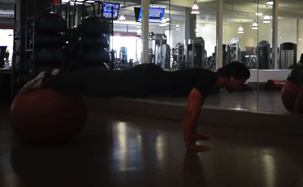 Bilde av personlig trener Cornelis Elander som utfører en push-up øvelse med føttene plassert på en stor treningsball (Swiss ball). Han er i en plankeposisjon med armene strake og hendene plassert på gulvet, mens føttene er balansert på ballen, noe som øker vanskelighetsgraden ved å engasjere kjernemuskulaturen mer. Mannen har på seg en mørk t-skjorte og mørke bukser. Treningsstudioet har speilvegger, og i bakgrunnen kan man se forskjellige treningsapparater og monitorer som viser TV-programmer eller treningsinformasjon. Denne øvelsen er effektiv for å styrke bryst-, skulder- og tricepsmuskulaturen, samtidig som kjernemuskulaturen jobber for å stabilisere kroppen på den ustabile ballen. Dette er en styrkeøvelse med fordeler for bein og rumpe, og en av styrkeøvelsene i vårt 30-dagers gratis treningsprogram og styrkeprogram.