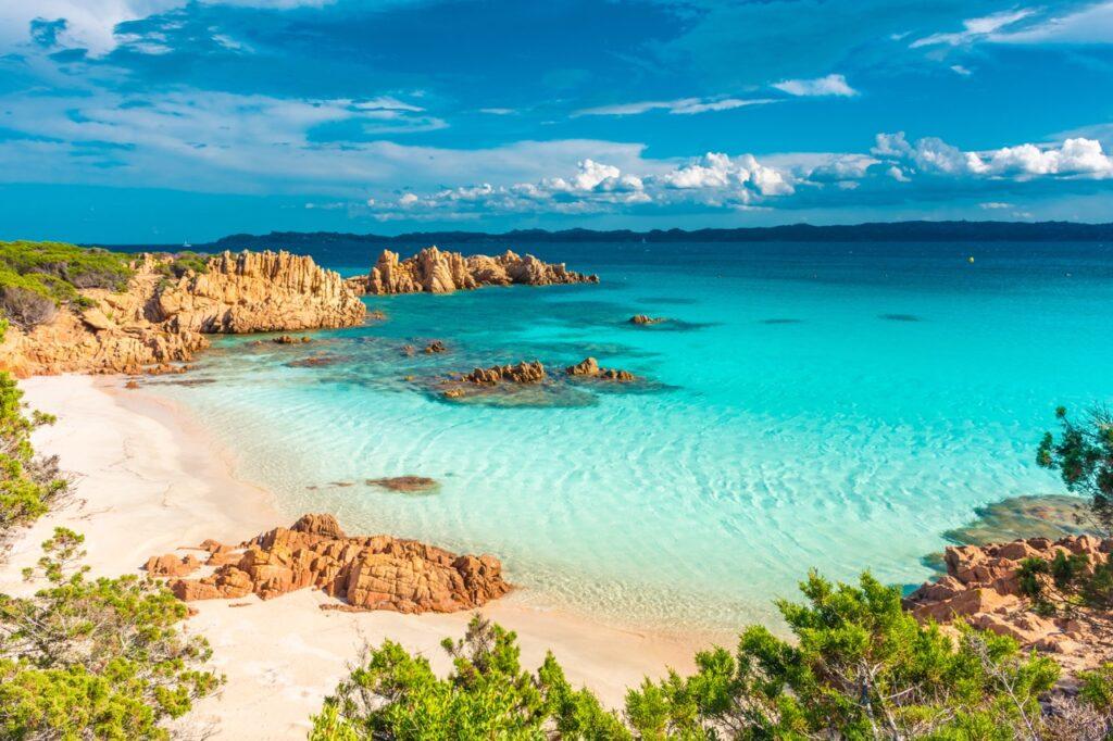 Et bilde av en vakker strand på Costa Smeralda, Sardinia, Italia. Stranden har hvit sand og krystallklart turkis vann som gradvis blir dypere. Klippeformasjoner stikker ut i vannet, og grønne busker og trær omgir stranden. I bakgrunnen strekker horisonten seg under en klar blå himmel med noen få hvite skyer. Det rolige havet skaper en idyllisk og avslappende atmosfære. Middelhavsøya Sardinia byr på både luksus og spennende historie, og her finner du krystallklar sjø og kritthvite strender. Vi gir deg tips til hva du kan se og gjøre på den italienske øya i Middelhavet; Dra til Smaragdkysten, Grotta di Nettuno, Nuraghe-ruinene, La Maddalena-øygruppen. Vi hjelper også med å finne hotell på Sardinia.