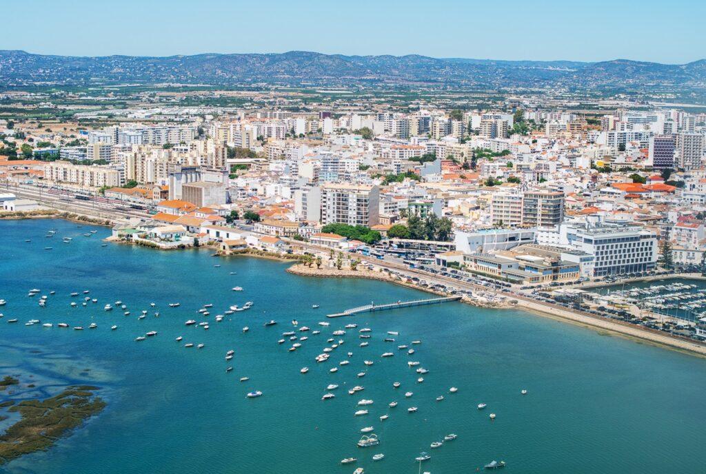 Bildet viser byen Faro langs Algarve-kysten i Portugal. Det er en oversikt over byen med mange bygninger, inkludert høye leilighetskomplekser og lavere bygninger nær vannkanten. Foran byen ligger en bred havn med mange små båter ankret opp i det blågrønne vannet. I bakgrunnen kan man se det landlige landskapet og åsene som omgir byen. Vi hjelper deg med å planlegge ferien til Algarve-kysten i Portugal. Vi gir deg alt fra tips til byer; Lagos, Faro, Tavira, Albufeira og Sagres. Samt hvor du kan finne hotell langs Algarve-kysten og shoppingmulighetene.