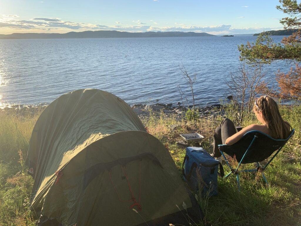 Person som sitter ved et grønt telt som er slått opp ved et vann i Norge. Solen skinner og det er fredfullt rundt plassen hvor teltet er slått opp. Vi har snakket med senior kommunikasjonsrådgiver i NAF, Nils Sødal, som forklarer hva du må tenke på før du velger å campe i det fri. Det finnes nemlig noen regler du må følge. Her får du reglene for fricamping på norgesferien.