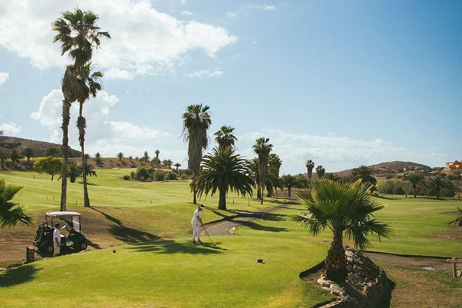 Bildet viser en golfbane med grønne fairways og palmer spredt rundt på området. I forgrunnen står en golfer som gjør seg klar til å slå en ball, mens en golfbil er parkert til venstre med en annen person ved siden av. Landskapet rundt golfbanen er kupert, og himmelen er blå med noen få skyer. Her får du ti ting du kan gjøre på Gran Canaria, Kanariøyene