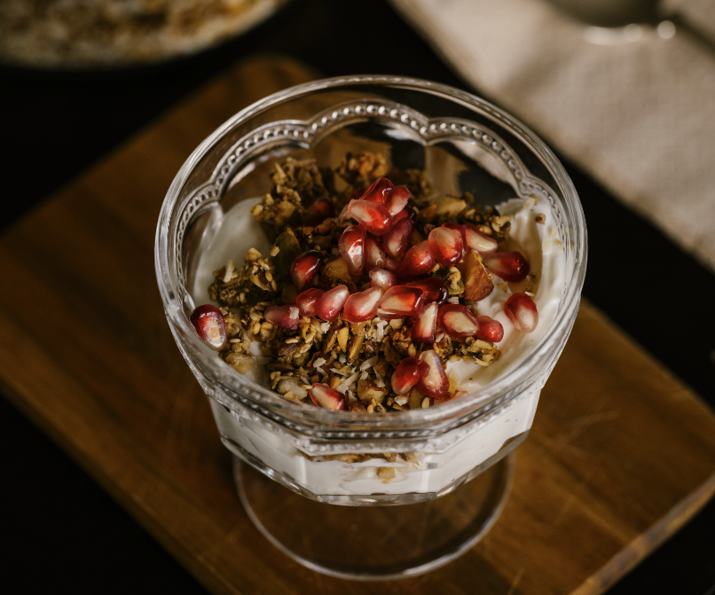 Bildet viser en glasskål med hjemmelaget granola. Skålen er fylt med en blanding av hvit yoghurt og sprø granola, og på toppen er det drysset med røde granateplekjerner. Granolaen ser ut til å inneholde ulike nøtter og frø, og det er en rustikk treplate under skålen. Bakgrunnen er noe uskarp, men det ser ut til å være en lys serviett eller klut i nærheten. Den delikate presentasjonen gir inntrykk av en sunn og appetittvekkende frokost eller snack. Vi har fått oppskrift på granola, en form for hjemmelaget og sunn ovnsbakt müsli av ernæringsfysiolog Ragnhild Lekven Fimreite. Forfatteren av boken 5 trinn til bedre fettforbrenning.