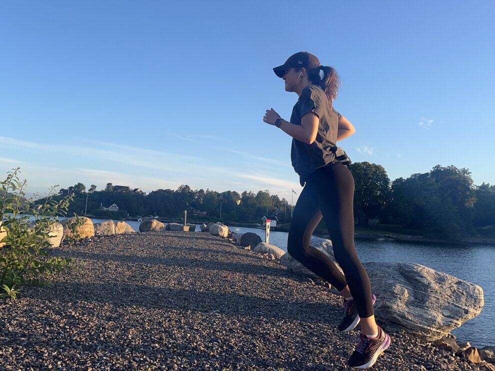 Bilde av en dame/kvinne som løper/ jogger ute på en pir som leder ut i Oslofjorden i naturen. I bakgrunnen ser man grusvein hun løper på, store steiner som omkranser piren, en øy, sjø og grønne, frodige trær. Kvinnen smiler og har på seg en NY Yankees-cpas, en svart tights, og en grå t-skjorte. Skoene er svarte med farge-innslag av lilla, hvitt, rød, og oransje og er løpesko som er best i test fra Brooks. Løpeekspert Andreas Gossner fra Løpetrening.no forteller hvilke muskler som brukes ved løping, hva som skjer med kroppen og gir gode tips på veien for deg som vil bli en god løper!