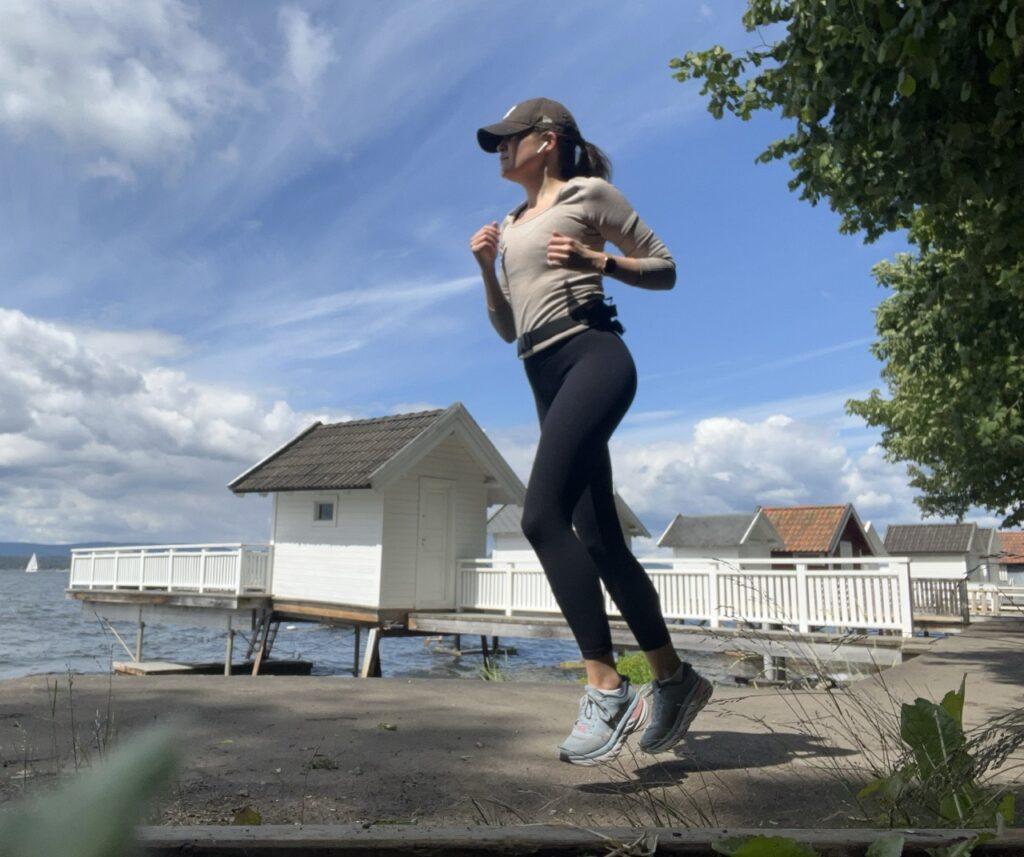 En kvinne som jogger ved sjøen. Hun har på seg en beige treningstop, svarte tights, og en svart caps. Hun smiler bredt mens hun løper, og har en smartklokke på håndleddet. Bakgrunnen viser en grønn plen med trær og en campingvogn. Solen skinner, og det er en lys og fin dag. Løpeekspert Andreas Gossner fra Løpetrening.no forteller hvordan du blir flink til å løpe, og gir deg treningsprogrammet for løping som du trenger!