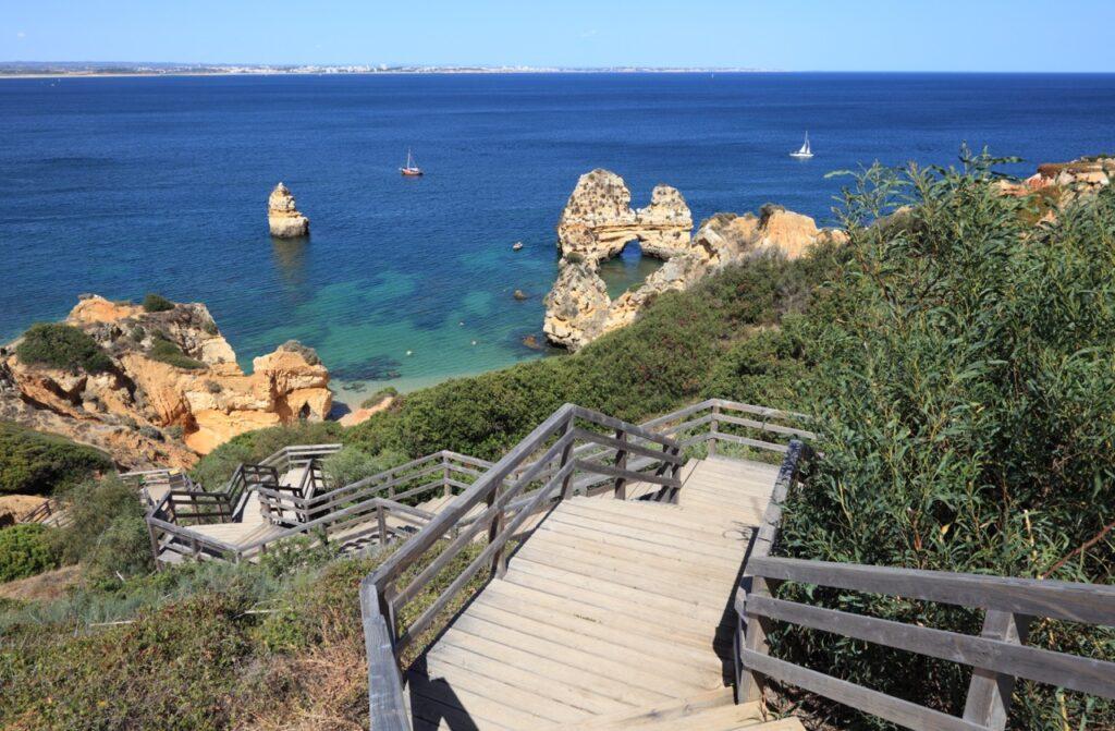 Bildet viser en naturskjønn utsikt over Lagos-kysten i Algarve, Portugal. En tretrapp med rekkverk slynger seg nedover en grønnkledd klippe mot en liten strand og det krystallklare, blågrønne vannet. I bakgrunnen stiger steinformasjoner dramatisk opp fra havet, og to seilbåter flyter rolig på vannet. Det klare været gir en vidstrakt utsikt over havet og kystlinjen i det fjerne. Vi hjelper deg med å planlegge ferien til Algarve-kysten i Portugal. Vi gir deg alt fra tips til byer; Lagos, Faro, Tavira, Albufeira og Sagres. Samt hvor du kan finne hotell langs Algarve-kysten og shoppingmulighetene.